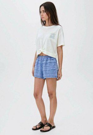 T-shirt blanche cintrée avec une poche graphique bleu clair, associée à un short bleu clair orné de dentelle et des sandales plates noires.