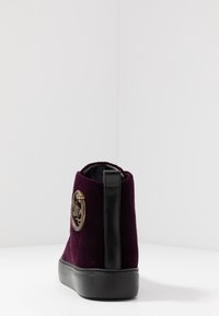 Fioletowe welurowe trampki za kostkę z czarną gumową podeszwą i zakładką na obcasie, z dekoracyjnym złotym emblematem po boku.