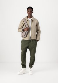 Beige jakke, hvit t-skjorte, olivengrønne bukser og hvite joggesko. Modellen bærer en lys grå skulderveske og står foran en hvit bakgrunn.