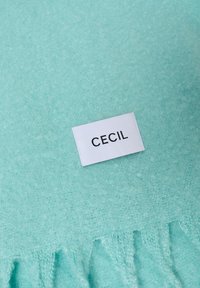 Jasno miętowy koc o miękkiej teksturze, z metką z napisem "CECIL" w czarnym kolorze, umieszczoną na białym prostokątnym tle.