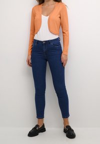 Mujer vestida con un cárdigan corto naranja sobre una camiseta blanca, combinado con unos jeans ajustados oscuros y mocasines negros, de pie contra una pared lisa.