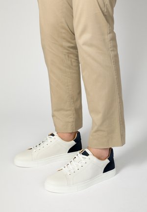 Białe skórzane sneakersy z granatowymi akcentami, płaska podeszwa i sznurowany design. Noszone z khaki spodniami, z zaokrąglonym czubkiem i minimalnymi detalami.