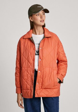 Giacca arancione trapuntata con bottoni a scatto, colletto e tasche frontali; indossata sopra una t-shirt bianca con grafica, abbinata a jeans scuri.