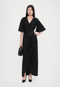 Woman wearing a black sequined wrap dress with wide sleeves, holding a black handbag, and wearing black pointed heels, standing against a white background.