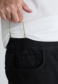 Pantalones negros con un acabado texturizado y una cinturilla elástica, que presentan un cordón verde que se extrae de una camiseta blanca que se lleva debajo.