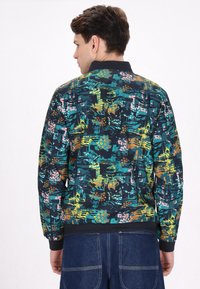 Jachetă bomber multicolorată cu un model geometric în nuanțe de teal, galben, portocaliu și roz; dispune de manșete ribbed și guler solid negru.