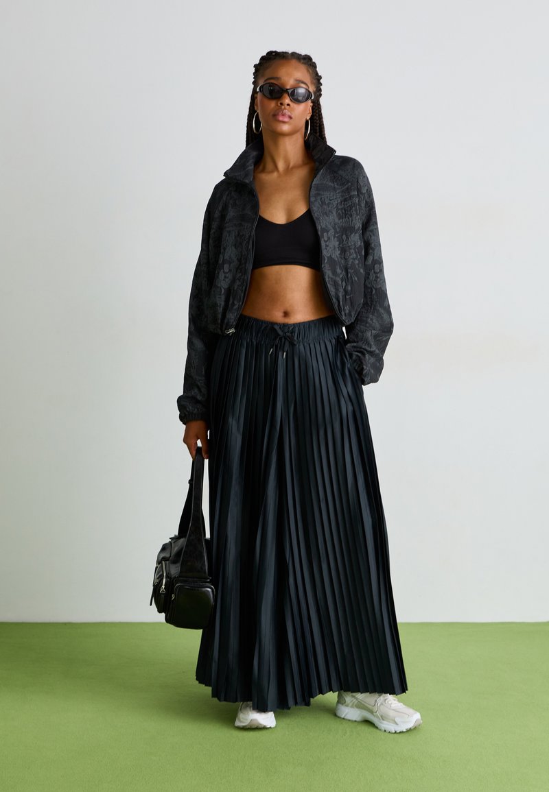 Chaqueta bomber negra con estampado floral, top corto negro, falda maxi plisada negra, zapatillas blancas y bolso negro, contra un fondo claro.