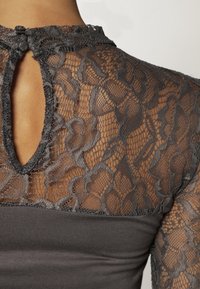 Grå topp med en blommig spetsdesign och en nyckelhålsöppning bak. Den släta tyget fram kontrasterar med den texturerade spetsen.