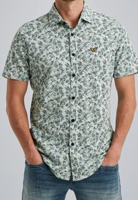 Camisa de manga corta con botones en verde claro, con un estampado de hojas en negro y verde oscuro. Incluye un pequeño logo bordado y botones negros.