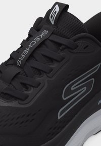 Κοντινό πλάνο μαύρου αθλητικού παπουτσιού Skechers που δείχνει τα κορδόνια, την ταμπέλα της γλώσσας με το λογότυπο, την υφή στην κορυφή και την άσπρη επένδυση στην άκρη της σόλας.