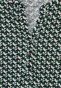 Blůza s vzorem obsahující geometrické tvary v odstínech modrozelené, bílé a černé. Má zářezový límeček a tři černé knoflíky po délce předního dílu.