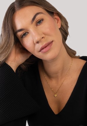 Young woman with light brown hair and blue eyes, wearing a black top and gold necklace, resting head on hand against plain background.