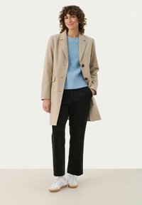 Beige Wollmantel mit schwarzen Knöpfen, hellblaue gerippte Sweater, schwarze Cordhosen und weiße Sneaker mit Gummisohle.