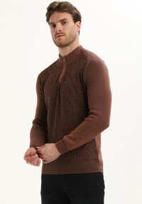 Brązowy sweter dzianinowy z wysokim kołnierzem i zamkiem, zdobiony geometrycznym wzorem na korpusie oraz ściągaczami przy rękawach, wykonany z miękkiego materiału.