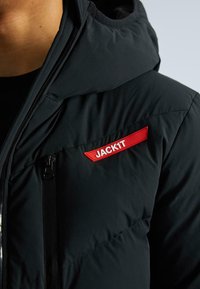 Artimas juodos paminkštintos striukės vaizdas su raudonu pleistru, pažymėtu "JACK1T", ant krūtinės šalia vertikalaus užtrauktuko kišenės.