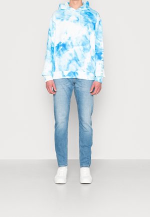 Sudadera azul tie-dye con acentos blancos, de tela de algodón, con capucha ajustable y cordón, combinada con jeans ajustados de color azul claro y zapatillas blancas.
