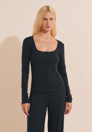 Blonde vrouw draagt een zwart lang mouw knooptop en bijpassende zwarte broek, staand tegen een beige achtergrond.