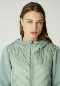 Lichtgroene zip-up jack met een gewatteerde voorkant en ribbelpanelen aan de zijkanten, met een capuchon en subtiele branding op de borst.