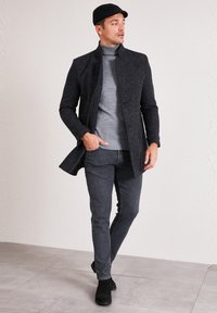 Cappotto nero texturizzato con colletto alto, dolcevita grigia, jeans grigio scuro e stivali neri, posizionati davanti a una parete chiara e un pavimento piastrellato.