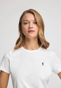 Vit bomull t-shirt med kort ärm, som har en liten broderad figur i mörkblått på vänster bröst. Enkel, klassisk design.