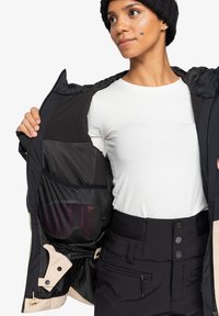 Svart isolert jakke med beige detaljer, med en mesh-innerlomme. Modellen har på seg en hvit skjorte og høyhælte svarte bukser.