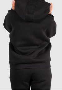 Personne portant un sweat à capuche noir et un pantalon noir, ajustant la capuche avec les deux mains, vue de dos contre un fond uni.