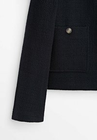 Musta teksturoitu takki, jossa on yksi rintatasku hopeanvärisellä napilla, suorat hihat ja hieman jäsennelty muotoilu.