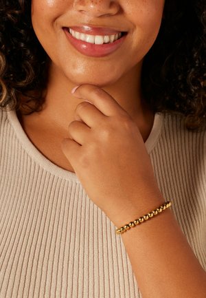 Gold bead bracelet on a wrist, featuring round beads and a smooth texture, paired with a light beige ribbed top.