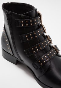 Bottines en cuir noir avec trois sangles, clous dorés, fermeture éclair sur le côté et un petit talon bloc. Texture lisse et design minimaliste.