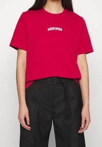 Röd T-shirt i bomull med korta ärmar och rund halsringning. Framsidan har en vit "WOOD WOOD" logotyp. Matchas med svarta byxor.