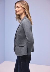 Femme aux cheveux blonds portant un blazer gris ajusté et un jean foncé, debout de profil devant un fond bleu clair.