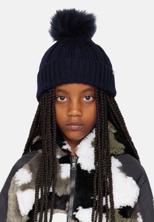 Beanie lavorato a maglia blu navy con un pompon in pelliccia sintetica rotondo in cima. Presenta un risvolto a coste piegato, indossato con una giacca in pile bianca e nera.