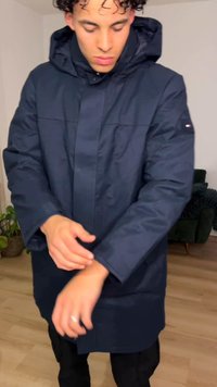 Nuori henkilö, jolla on kiharat hiukset, säätää tummansinisen hupullisen talvitakin hihaa olohuoneessa.