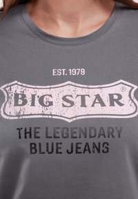 Pilkas medvilnės marškinėliai su dideliu rožiniu ir juodu grafiniu logotipu, kuriame yra tekstas "BIG STAR EST. 1979 LEGENDINIAI MĖLYNI DŽINSAI."