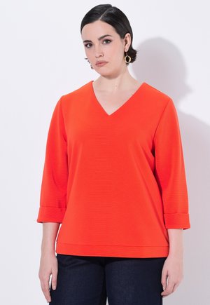 Oranje V-hals top met driekwartmouwen en opgerolde manchet, gecombineerd met donkere denim jeans. Glad textuur, geen patronen zichtbaar.