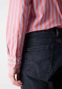 Camisa de mangas largas a rayas rosas y azules con puños abotonados, combinada con jeans de mezclilla oscuros que presentan un detalle en la costura de los bolsillos.
