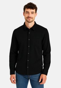 Mann in schwarzem Hemd mit Knöpfen und Brusttasche sowie blauen Jeans, der vor einem schlichten hellen Hintergrund steht.