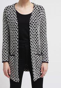 Svart och vit mönstrad cardigan med lös passform, öppen front och långa ärmar. Har en texturerad stickning och sidofickor.