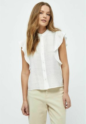 Weiße Bluse mit kurzem Ärmel und Rüschendetails, mit Knopfleiste vorne und aus leichtem, strukturiertem Stoff. Kombiniert mit hellbeigen Hosen.