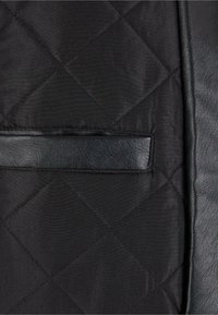 Veste matelassée noire avec un motif diagonal, rehaussée de cuir noir lisse sur les côtés et d'une petite poche à l'avant.