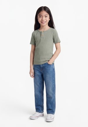 Young girl smiling, standing with one hand in pocket, wearing green short-sleeve shirt, blue jeans, and white sneakers on white background.