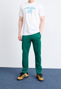 Vit t-shirt med blå text och Adidas-logotyp, kombinerad med gröna träningsbyxor med sidostråk, samt bruna sneakers med svarta ränder.