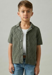 Zelený krátky cardigan s krátkymi rukávmi a diamantovým pleteným vzorom, s golierovým výstrihom a zapínaním na gombíky, nosený cez bielu tielko.