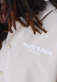 Chaqueta beige de botones con una textura suave. Presenta texto blanco "PROJECT X PARIS" y "MEMBERS' CLUB" en el lado izquierdo.