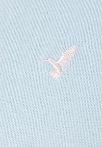 Tessuto a maglia azzurro chiaro con un piccolo logo di uccello ricamato in bianco. La texture è liscia con un sottile motivo a coste.