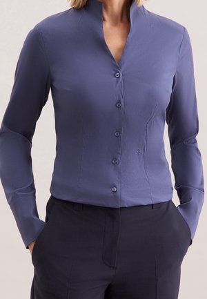 Woman wearing a fitted, long-sleeve blue button-up shirt tucked into dark trousers with hands in pockets against a neutral background.