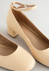Beige texturierte Blockabsatzschuhe mit runder Zehenform und einem Knöchelriemen. Das Material hat ein gewebtes Muster. Glatte Innenauskleidung.