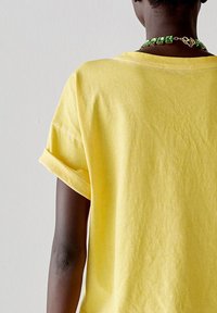 T-shirt giallo in cotone con maniche arrotolate, texture liscia e scollatura rotonda. È indossata una collana di gioielli verde.