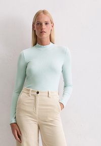 Hellblaues langärmliges Oberteil mit einem Mock-Neck, kombiniert mit cremefarbener Cordhose mit hoch sitzendem Bund und vertikalen Noppen sowie einem einzigen Knopfverschluss.