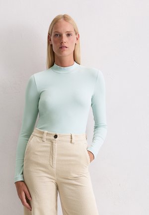 Maglietta azzurro chiaro a maniche lunghe con colletto alto, abbinata a pantaloni in corduroy beige a vita alta con righe verticali e chiusura a un bottone.